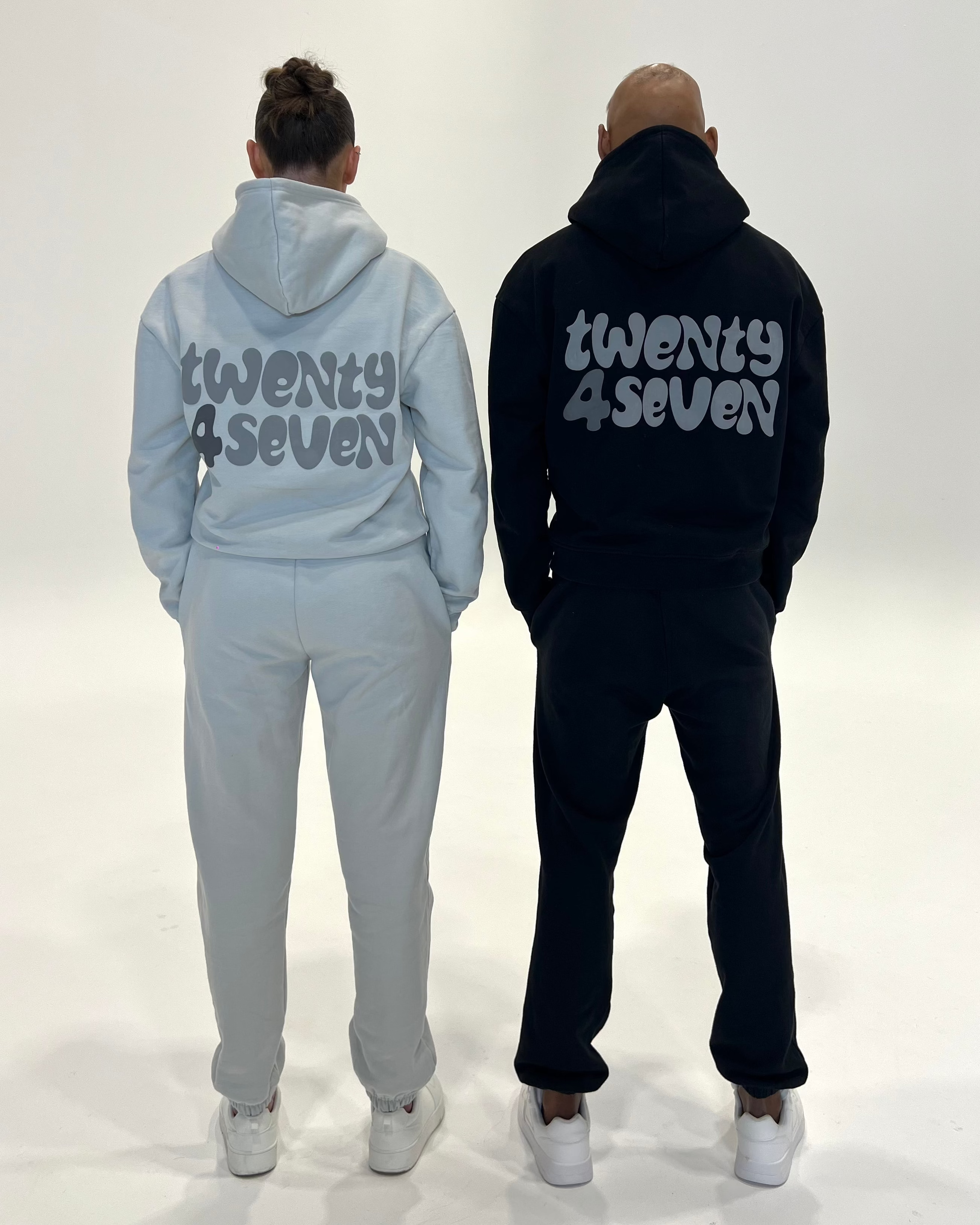 Two models standing from the back wearing matching sweatshirt and jogger sets in light grey and black, both featuring the text 'twenty 4 seven' printed in a stylized font on the back.