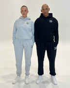 Two models standing side-by-side in matching tracksuits, one in a light grey set and the other in a black set, both featuring a white '24' graphic on the chest and thigh, photographed against a seamless white studio background.